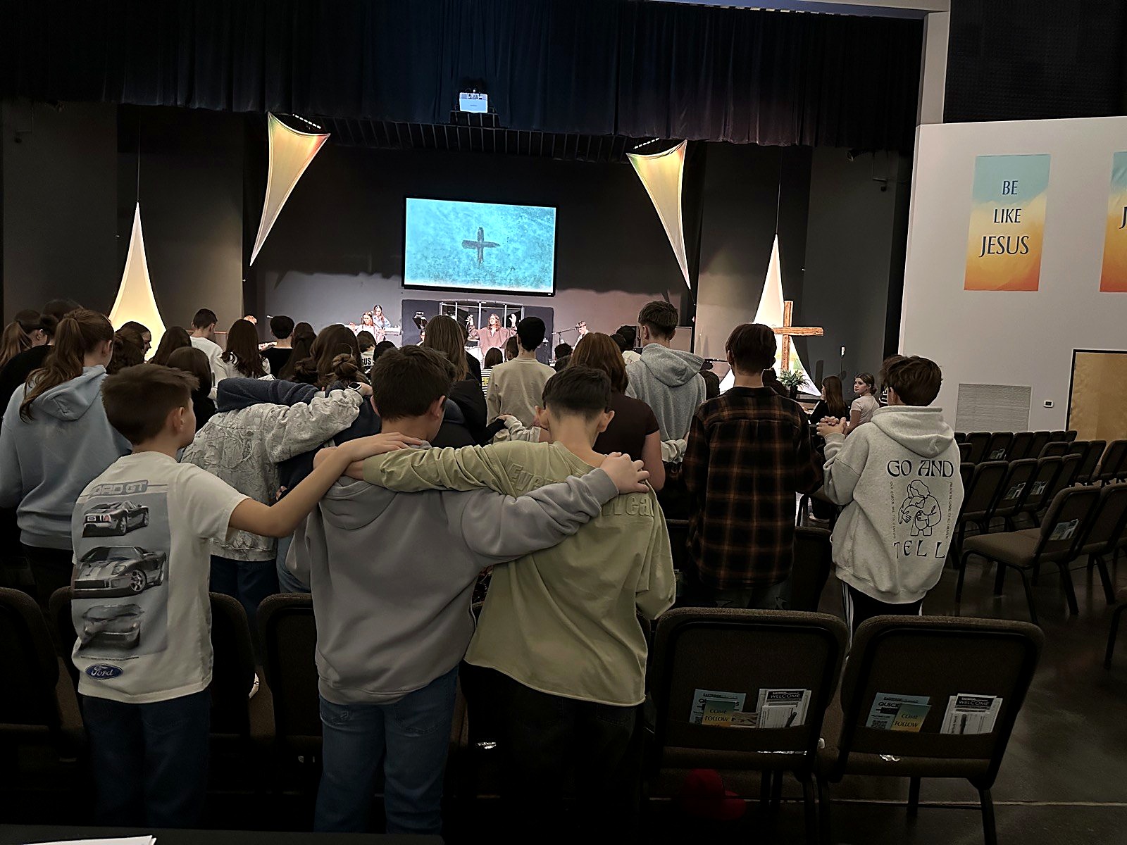 Spring Mountain students standing arm in arm in worship during chapel, facing a cross and a Be Like Jesus banner